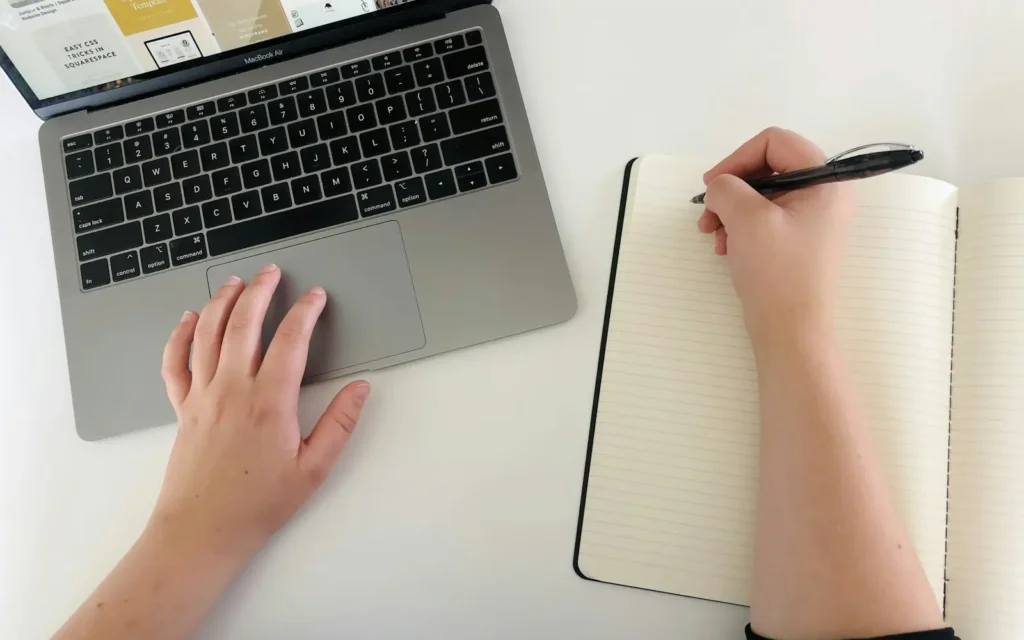 A learner writing notes beside a laptop
